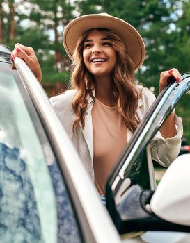 A cute woman in a hat stands near the car on the background of the forest. Purchase, car rental. Rest in nature, weekend.