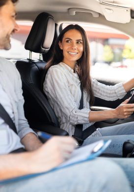 Happy pretty young woman sitting inside car with man instructor by her side, lady student holding hands on steering wheel, passing exams successfully at driving school, getting licence